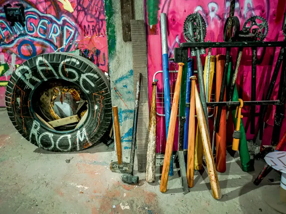 Tacos e marretas encostados na parede grafitada da sala, ao lado de um pneu pintado com “Rage Room”.