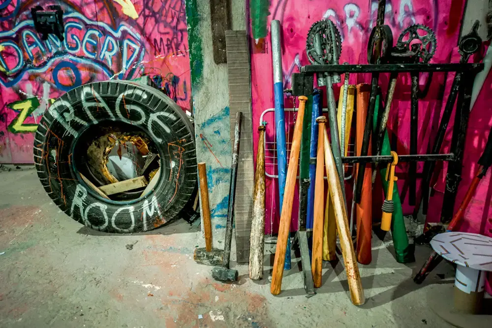 Tacos e marretas encostados na parede grafitada da sala, ao lado de um pneu pintado com “Rage Room”.